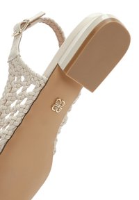 Woven white sandal with a flat sole, ankle strap, and a small metallic logo detail on the footbed; smooth texture and open design.