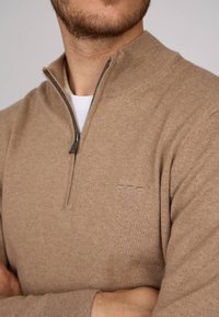 Pull à zip marron avec un col montant, confectionné en maille douce. Présente un logo discret sur la poitrine et des poignets côtelés.