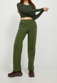Frau, die ihre Arme streckt, trägt ein dunkelgrünes, langärmliges Crop-Top, olivgrüne JJXX-Hosen und braune Sneaker, steht vor einem einfarbigen Hintergrund.