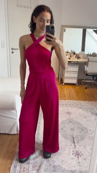Fuchsia jumpsuit with v-neck, cross-back design, wide legs, and a smooth texture. Worn with black shoes on a patterned rug.