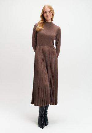 Długa, brązowa dzianinowa sukienka z wysokim kołnierzem, długimi rękawami i plisowanym dołem. Stylizowana z czarnymi, wysokimi na kolana botkami. Gładka tekstura, ciepły ton.
