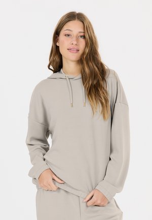 Ung kvinde med langt, bølget hår iført en lys beige hættetrøje og matchende bukser, stående mod en ensartet hvid baggrund.