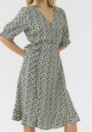 Wiązana sukienka kopertowa w kwiaty na zielonym tle z różowymi i białymi kwiatami, dekolt w kształcie litery V, krótkie bufiaste rękawy oraz wiązanie w talii.
