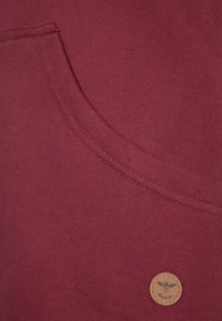 Bordowy materiał bluzy z kapturem o gładkiej fakturze, z przednią kieszenią oraz małą, okrągłą skórzaną metką z wytłoczonym wzorem pszczoły.