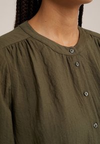 Olijfgroene knoopshirt met een ronde hals, gekreukt textuur en subtiele plooien bij de schouders; vijf knopen zichtbaar.