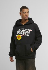 Sort hættetrøje med en frontgrafik, der har "Enjoy Coca-Cola" i hvid samt en gul tegneserie-stil "YES!" boble. Der er en kengurulomme inkluderet.