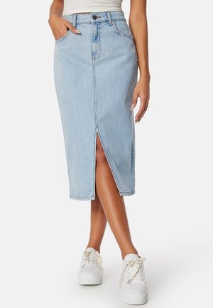 Lyseblå denim midinederdel med frontslids, båret med hvide sneakers og en hvid top stoppet ned, modellens venstre hånd i lommen.