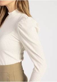 Vit texturerad långärmad topp med hög halsringning och puffärmar. Nedanför syns en figursydd khaki-plagg delvis.