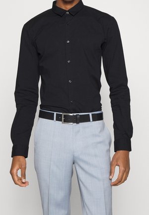 Homme portant une chemise noire ajustée boutonnée rentrée dans un pantalon de costume gris clair avec une ceinture noire.