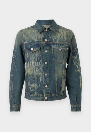 Młody mężczyzna w dżinsowej kurtce z nadrukami, brązowej bluzie z kapturem, kremowych luźnych spodniach i brązowych butach roboczych, ręce w kieszeniach.