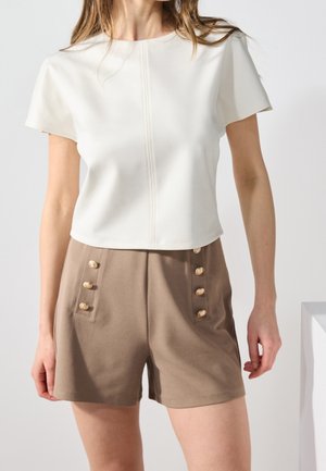 Kvinde iført en hvid kortærmet top og high-waist taupe shorts med to rækker af gyldne knapper på frontlommerne.