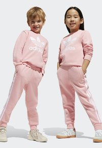 Ružové bavlnené mikiny a nohavice s bielym logom Adidas a pruhmi. Textúrovaná látka; pružné manžety; ľahké topánky v béžovej a zelenej farbe.