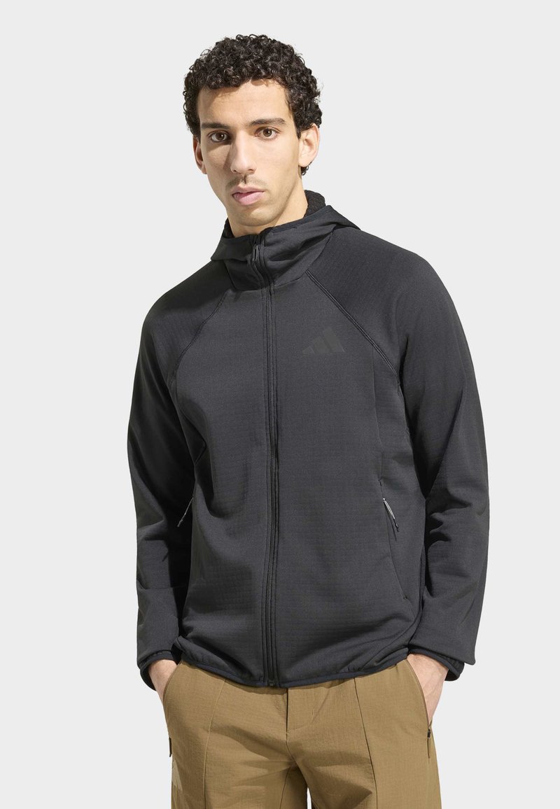 Jeune homme aux cheveux bouclés portant un sweat à capuche noir zippé et un pantalon beige, debout les mains dans les poches devant un fond uni.