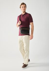 Sac en bandoulière en cuir noir avec des accents marron, poche zippée et bandoulière réglable, porté par un homme en polo bordeaux et pantalons crème.