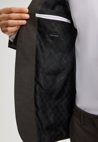 Giacca in lana marrone con fodera nera a fantasia. Presenta una tasca sul petto con bordo bianco e un'etichetta che dice "STRETCH WOOL."