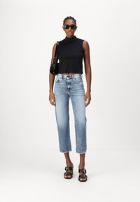 Top nero senza maniche, jeans azzurri a vita alta, sandali neri con tacco. La modella indossa occhiali da sole e si trova di fronte a uno sfondo neutro.