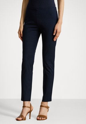 Vrouw draagt nauwsluitende donkerblauwe enkelhoge broek en bruine sandalen met hoge hak en enkelbandjes, staand op een effen lichte achtergrond.