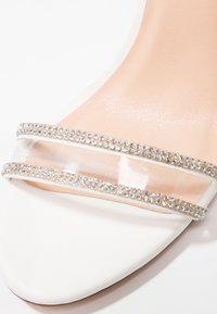 Sandale à talons hauts blanche avec une bride transparente ornée de strass argentés, texture de cuir lisse et design à bout pointu.