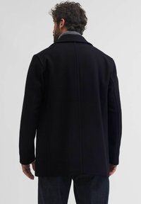 Melna vilnas auduma mētelis ar klasisku apkakli, iekļaujot teksturētu iekšējo apkakli un pielāgotu piegriezumu. Redzami šuvi un garas piedurknes.