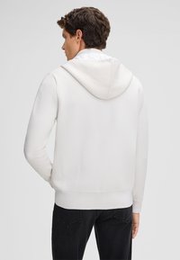 Weißer, strukturierter Hoodie mit Rückansicht, ausgestattet mit Kapuze und gerippten Bündchen, kombiniert mit dunklen Jeans für den Kontrast.