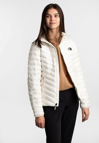 Junge Frau in einer weißen Steppjacke, einem beige Pullover und schwarzen Hosen, die vor einem schlichten weißen Hintergrund steht.