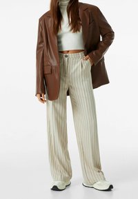 Brązowa oversize marynarka z imitacji skóry, biały prążkowany krótki golf, beżowe spodnie w szerokie prążki oraz kremowe sneakersy.