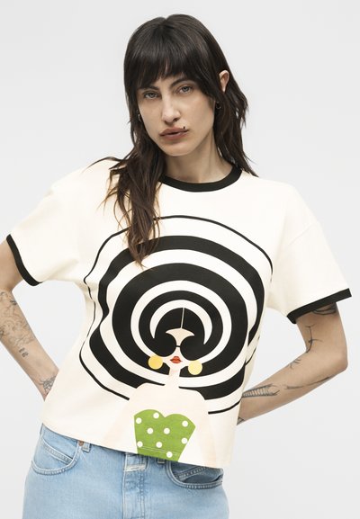 Femeie cu păr închis la culoare și tatuaje poartă un tricou alb cu un spiral negru mare și o femeie stilizată cu bluză verde cu buline și cercei aurii.