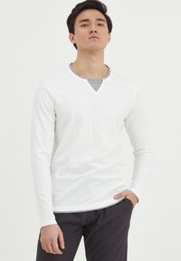 Hvid langærmet T-shirt med V-hals og gråt indvendigt foer, parret med mørke bukser. Glat stof, casual pasform, minimalistisk design.