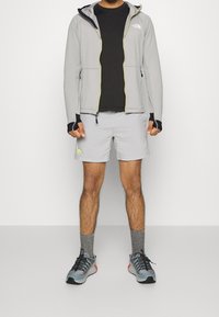 Chaqueta de cremallera gris claro con capucha, capa base negra, pantalones cortos gris claro y zapatillas gris-azul con acentos en negro y rojo.
