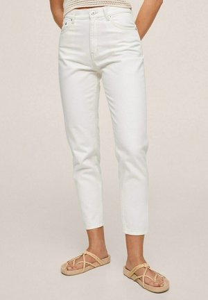 Person trägt hoch taillierte, weiße, eng anliegende Hose und beige Sandalen mit geflochtenem Riemen und steht vor einem schlichten, hellen Hintergrund.