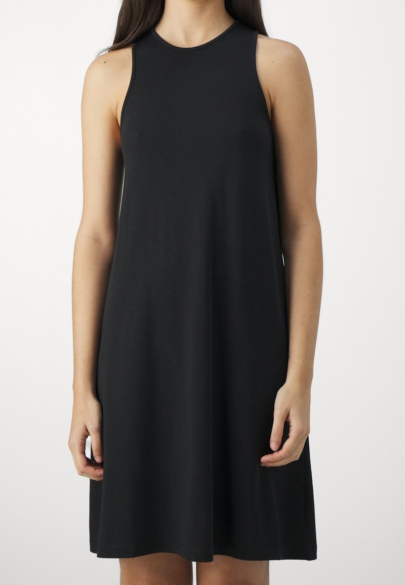 Nainen, joka on pukeutunut hihattomaan, mustaan, polveen ulottuvaan A-linjaisiin mekkoon, seisoo tasaisella valkoisella taustalla.