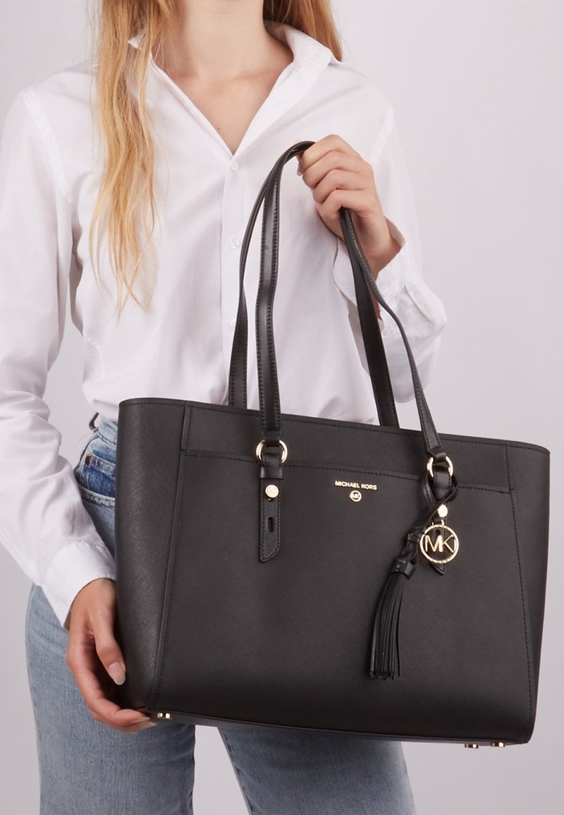 Femme en chemise blanche et jean tenant un sac cabas noir Michael Kors avec des doubles poignées et un porte-bonheur logo doré.