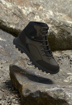 Bruine en zwarte wandelschoen met veters zwevend boven rotsachtige grond, met een robuuste zool en een achtergrond van buitengebied.