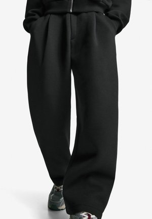 Person trägt lockere schwarze Hose und graue Sneakers mit Schnürsenkeln, steht mit den Händen in den Taschen vor einem weißen Hintergrund.
