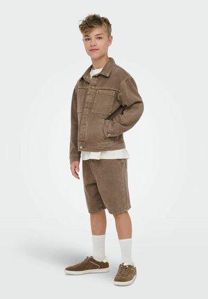 Ung person står, iført brun denimjakke og matchende shorts, hvite ankelsokker og brune joggesko, ser fremover.