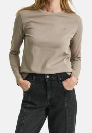 Frau trägt ein beiges Langarmshirt, das in hoch taillierte schwarze Jeans gesteckt ist, mit den Händen in den Taschen, stehend vor weißem Hintergrund.