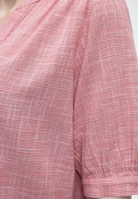 Blouse à carreaux rouges et blancs avec des manches courtes bouffantes et un ourlet à revers, mettant en valeur le tissu texturé et les détails des coutures aux épaules.