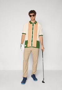 Kortærmet skjorte med orange og grønne lodrette striber, med en grøn krave og bunde accenter. Neutral beige bukser og golfske.