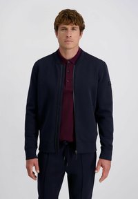 Marine blauwe rits-sweater met een gladde textuur en geribbelde manchetten, gecombineerd met een bordeauxrood poloshirt met drie knopen.