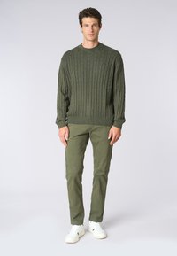 Maglione verde oliva a lavorazione a treccia con collo a costine, abbinato a pantaloni verde oliva e sneakers bianche. Il modello è in piedi contro uno sfondo neutro.