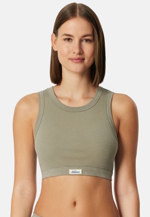 Mladá žena s rovnými hnědými vlasy, oblečená v tílku z žebrované látky světle olivové barvy, s malým logem vpředu.