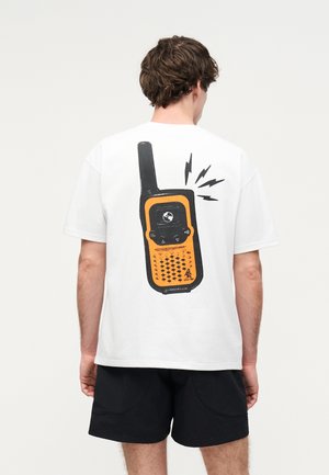 Mężczyzna noszący białą koszulkę z dużą pomarańczowo-czarną grafiką krótkofalówki na plecach, połączoną z czarnymi szortami.