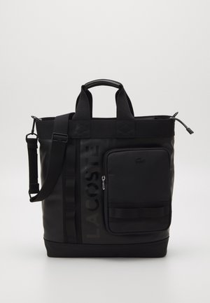 Bolso tote negro de Lacoste con asas dobles, correa de hombro ajustable, bolsillo delantero con cremallera y logo vertical en relieve sobre un fondo liso.