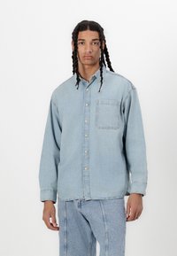 Cămășă din denim albastru deschis, cu butoni, dotată cu un buzunar frontal, mâneci lungi și un croi lejer, asortată cu pantaloni din denim albastru deschis.