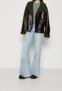 Veste en simili cuir noir avec col en polaire vert olive et poches, portée sur une chemise rayée et un jean évasé en bleu clair, avec des chaussures noires.