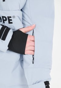 Vaaleansininen puffatakki mustilla yksityiskohdilla. Varustettu vetoketjullisella taskulla ja tiukasti istuvalla mustalla hihalla. Sileä pinta. Suuri logo esillä.