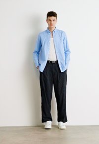 Camicia azzurra chiara con bottoni sopra una canotta bianca, abbinata a pantaloni neri a righe e sneakers bianche, in piedi su uno sfondo neutro.