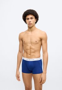 Μπλε βαμβακερά μποξεράκια με λευκή ζώνη μέσης που φέρει το λογότυπο Tommy Hilfiger, με εφαρμοστή γραμμή και ομαλή υφή. Το μοντέλο στέκεται με τα χέρια χαλαρά.