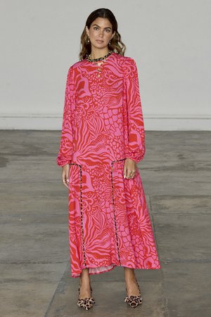 Long-sleeve, pink dress with bold, abstract patterns. Features a loose fit, rounded neckline, and decorative black trim. Paired with leopard print heels.