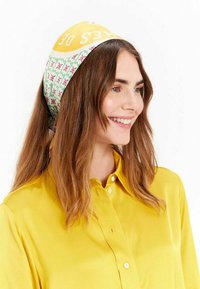 Foulard en soie avec une section jaune portant du texte blanc, entourée d'un motif floral vert. Porté noué à l'arrière.